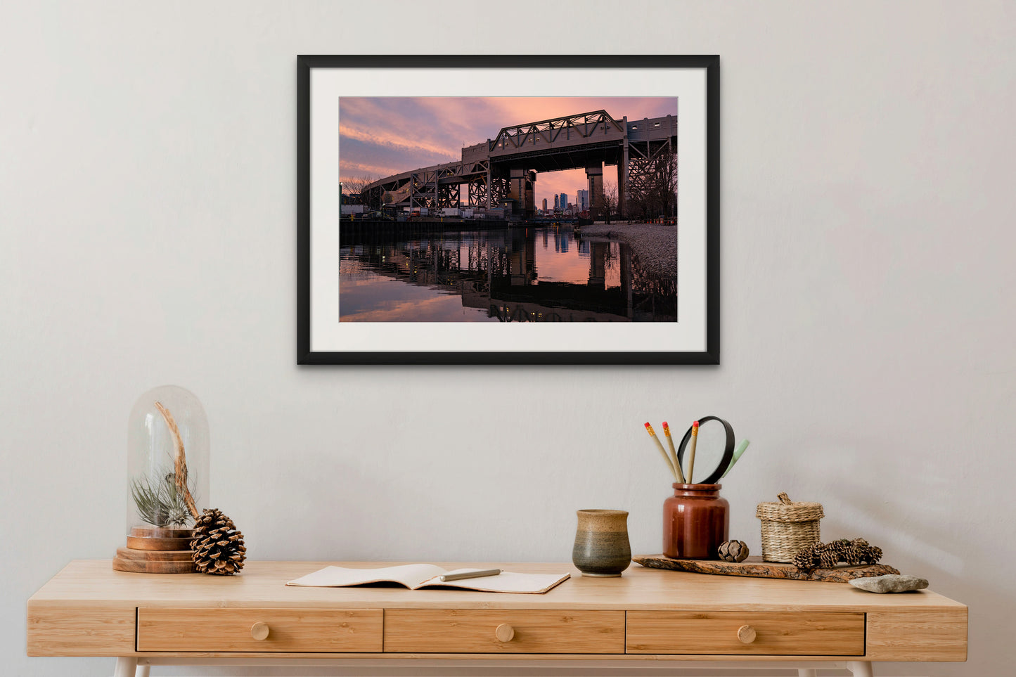 A framed print of sunset behind the Smith & 9th St. subway station viaduct in Gowanus, Brooklyn is hung above a desk with pine cones and an open notebook.